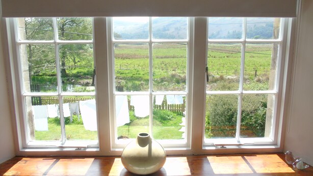 Une fenêtre donnant sur un paysage verdoyant, un vase blanc posé sur le rebord et un banc en bois.