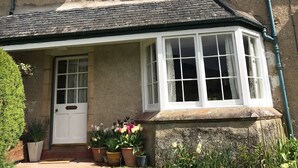 Une maison à deux étages, dotée d’un toit à pignon, d’une cheminée et d’un porche à l’avant, agrémenté de plantes en pot.