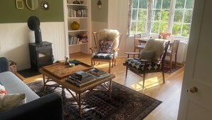 Un salon chaleureux avec une cheminée, une bibliothèque, un coin repas avec une table et des chaises, et une grande fenêtre avec des rideaux.
