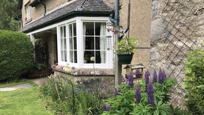 Une maison traditionnelle dotée d’une baie vitrée, entourée d’un jardin bien entretenu où poussent diverses plantes et fleurs.