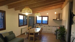 En hyggelig stue med træloft, spiseplads med bord og stole, samt et køkken med hvide skabe og udsigt ud af vinduerne.