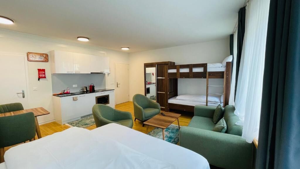 A compact hotel room with a bunk bed, a small kitchenette, a dining table, and two armchairs.
