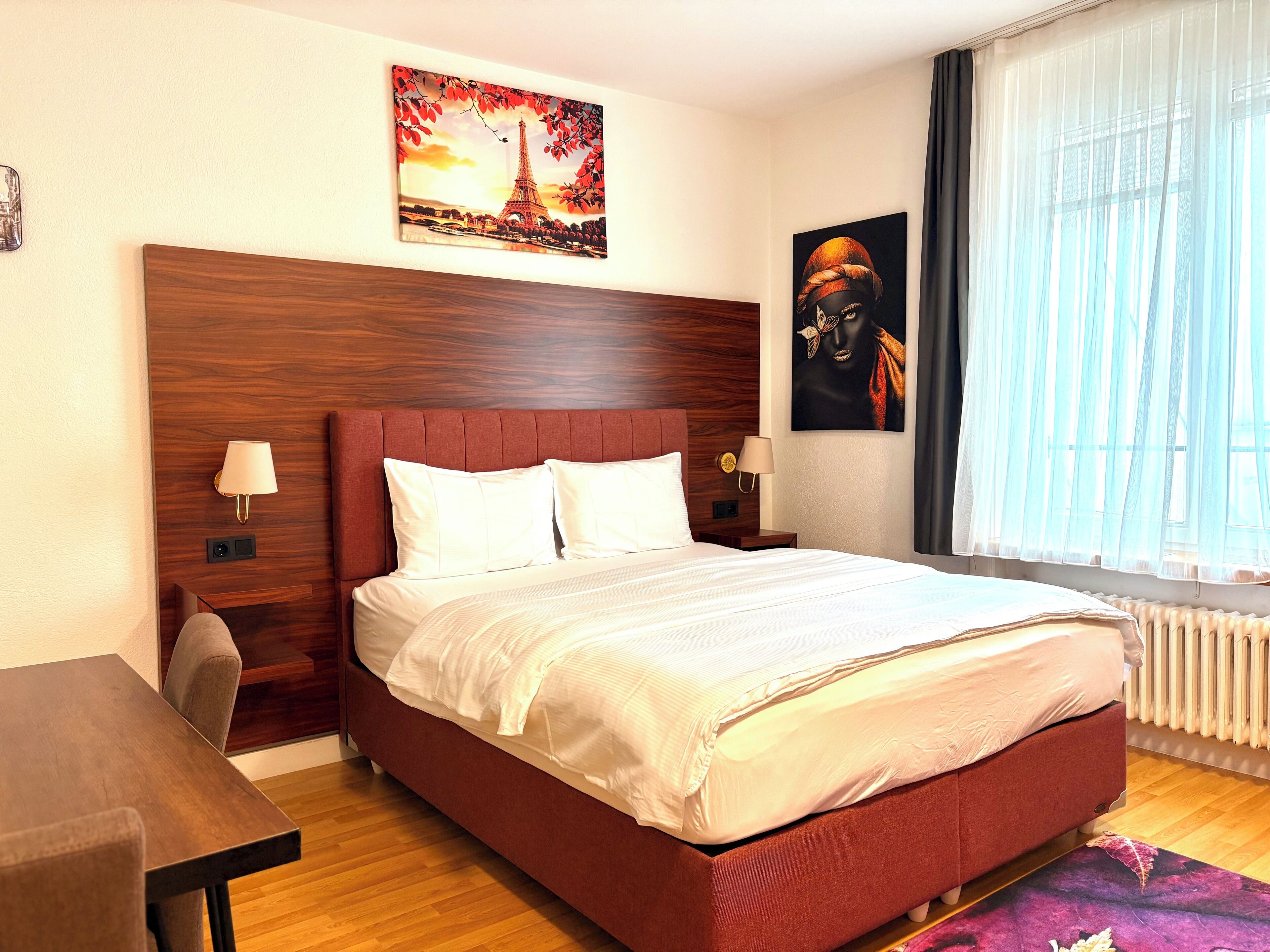 Ein Hotelzimmer mit einem großen Bett, zwei Nachttischen, einem Schreibtisch und einem Fenster mit durchsichtigen Vorhängen.