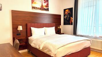 Ein Hotelzimmer mit einem großen Bett, zwei Nachttischen, einem Schreibtisch und einem Fenster mit durchsichtigen Vorhängen.