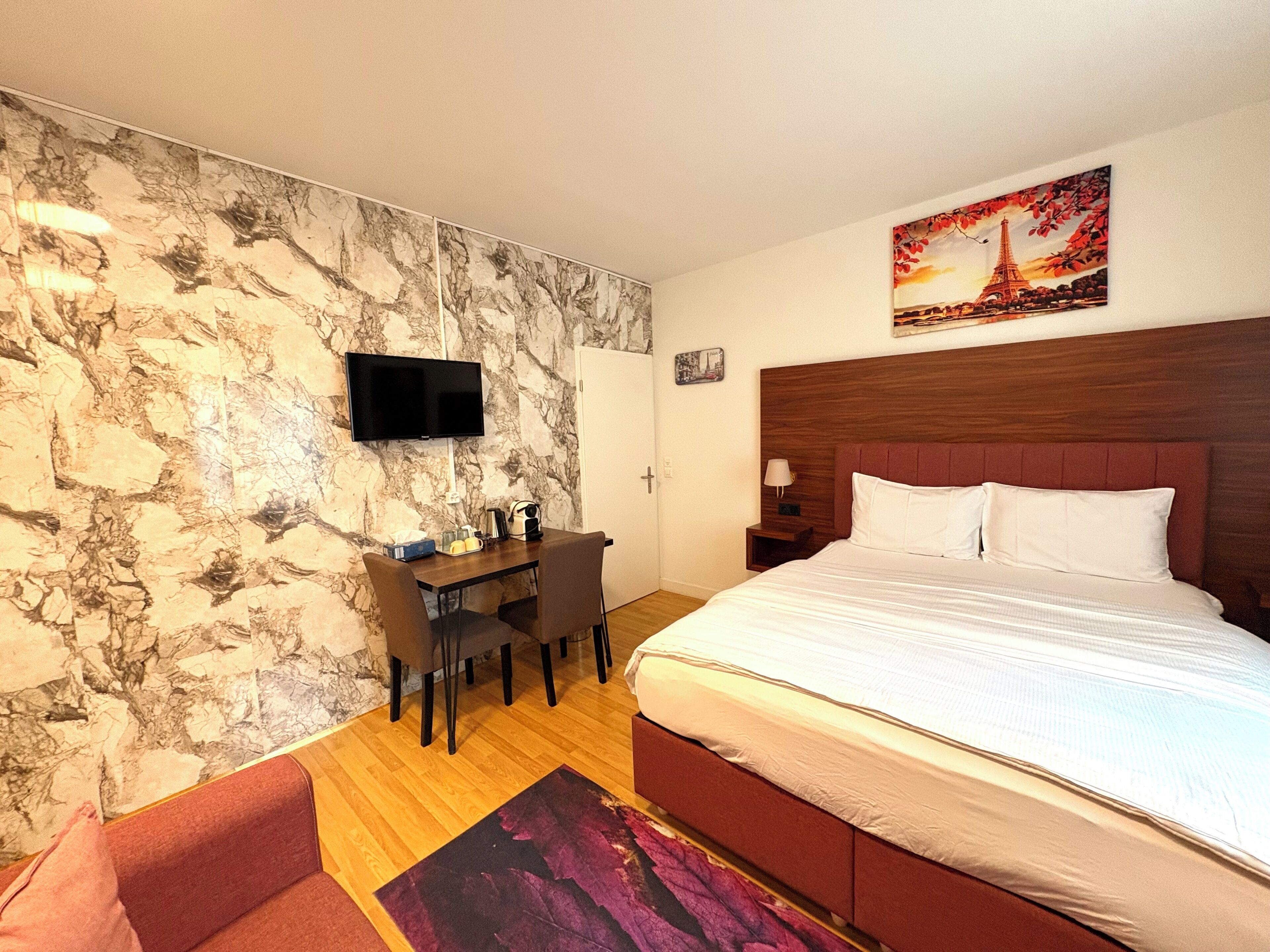 Ein Hotelzimmer mit Marmorwand, einem Bett mit weißen Leinentextilien, einem hölzernen Kopfteil, einem Schreibtisch mit Stuhl, einem an der Wand befestigten Fernseher und einem Gemälde vom Eiffelturm.