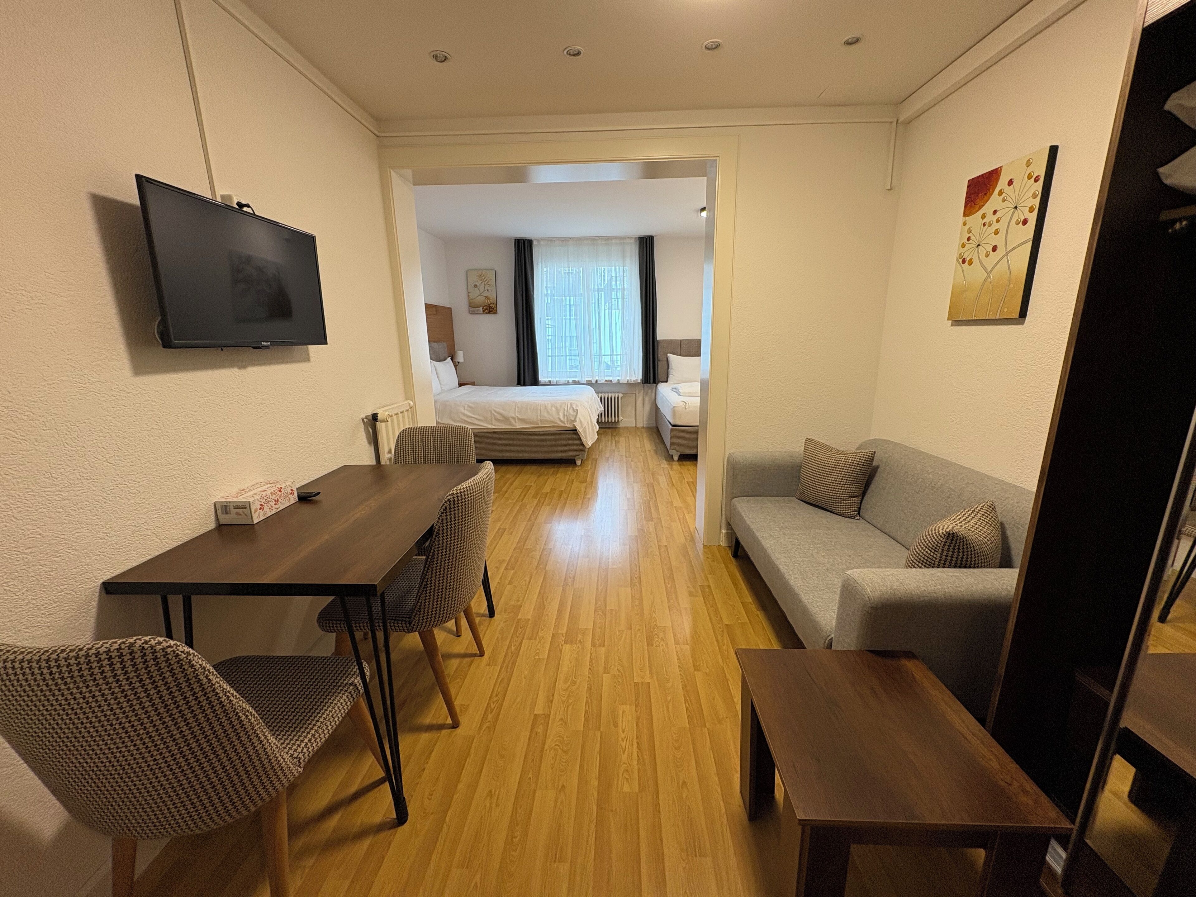 Una camera d'albergo compatta con pavimento in legno, un letto, un divano, una TV e un'area pranzo con sedie.
