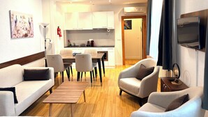 Una sala moderna con sofá, sillones, una mesita de centro, televisor y una cocina al fondo.