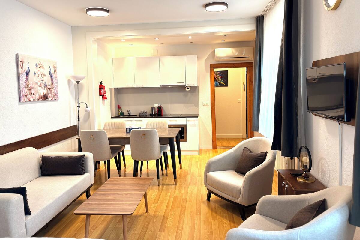 Ein modernes Wohnzimmer mit einer Couch, Sesseln, einem Couchtisch, einem Fernseher und einer Küchenzeile im Hintergrund.