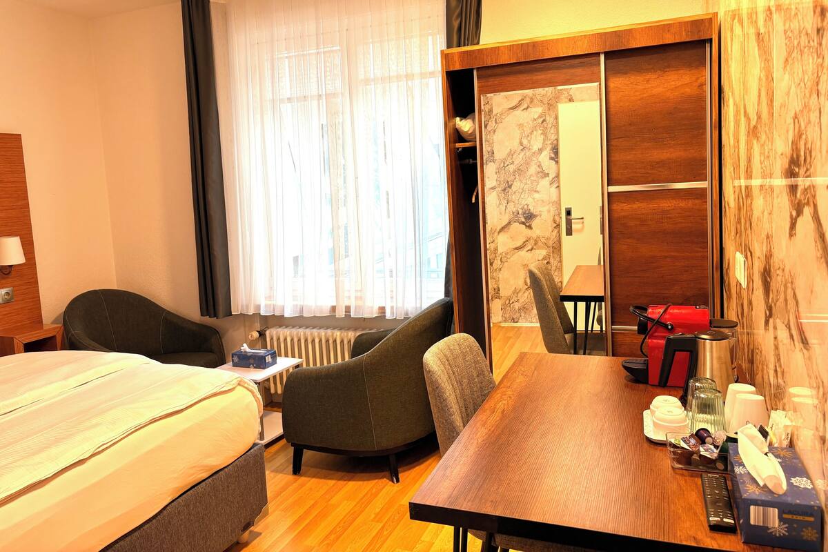 Ein Hotelzimmer mit zwei Betten, einem Esstisch, einem Stuhl, einem Heizkörper, einem Kleiderschrank und einem Fenster mit Vorhängen.