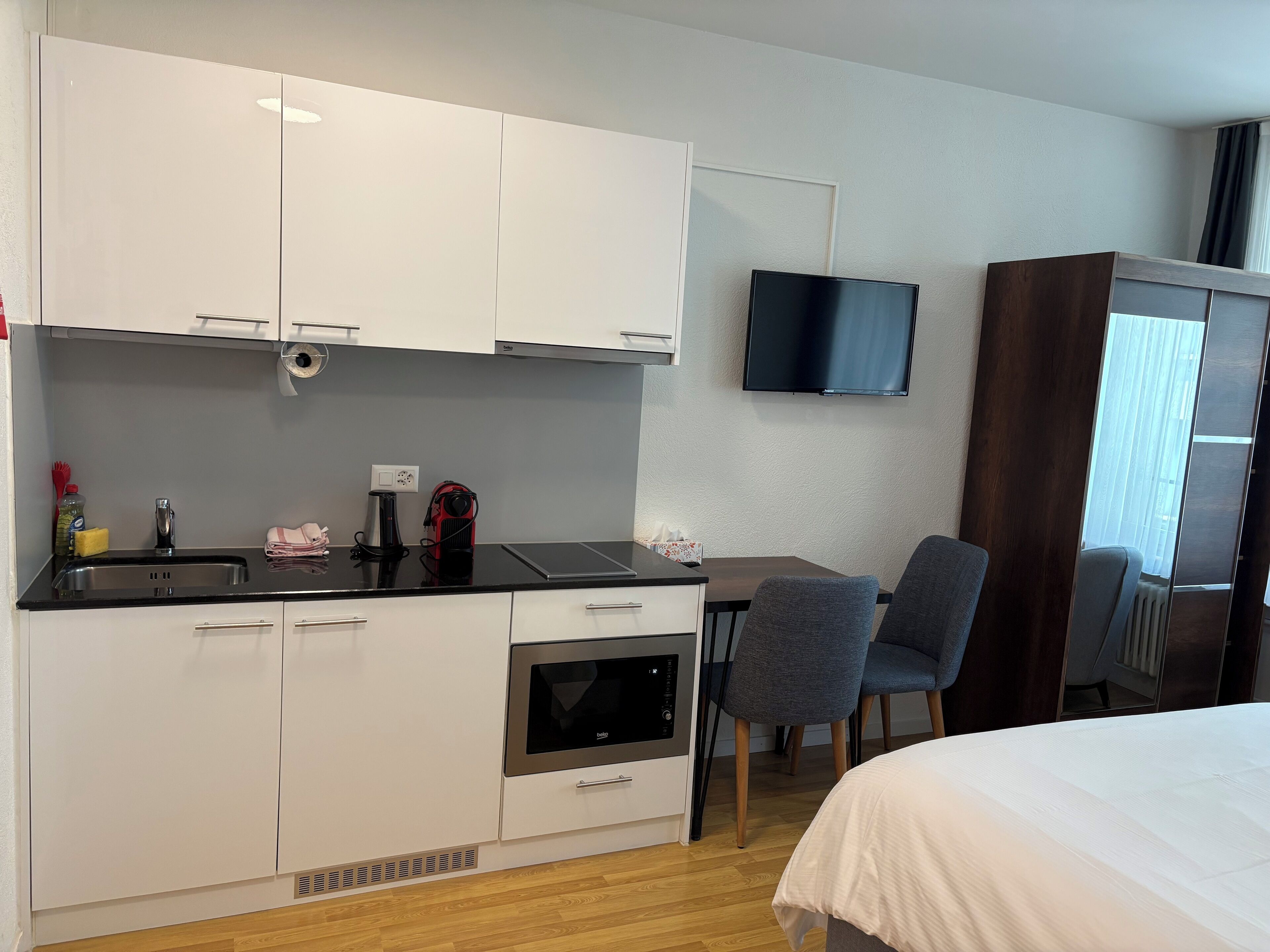 A compact hotel room with a kitchenette, a flat-screen TV mounted on the wall, a bed with a white duvet, and a window with curtains.