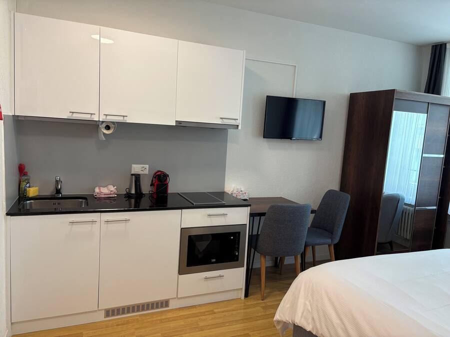 A compact hotel room with a kitchenette, a flat-screen TV mounted on the wall, a bed with a white duvet, and a window with curtains.