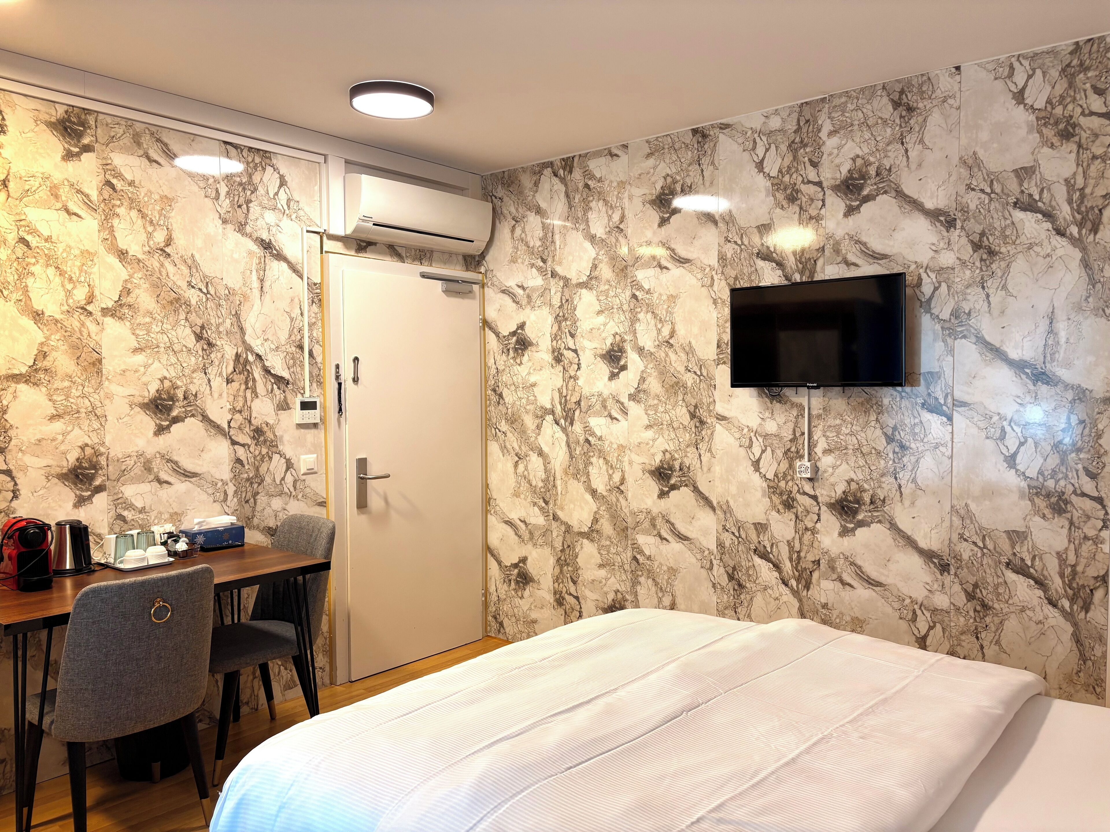Une chambre d’hôtel comprenant un lit, un bureau avec une chaise, un appareil de climatisation, un téléviseur fixé au mur et une porte.