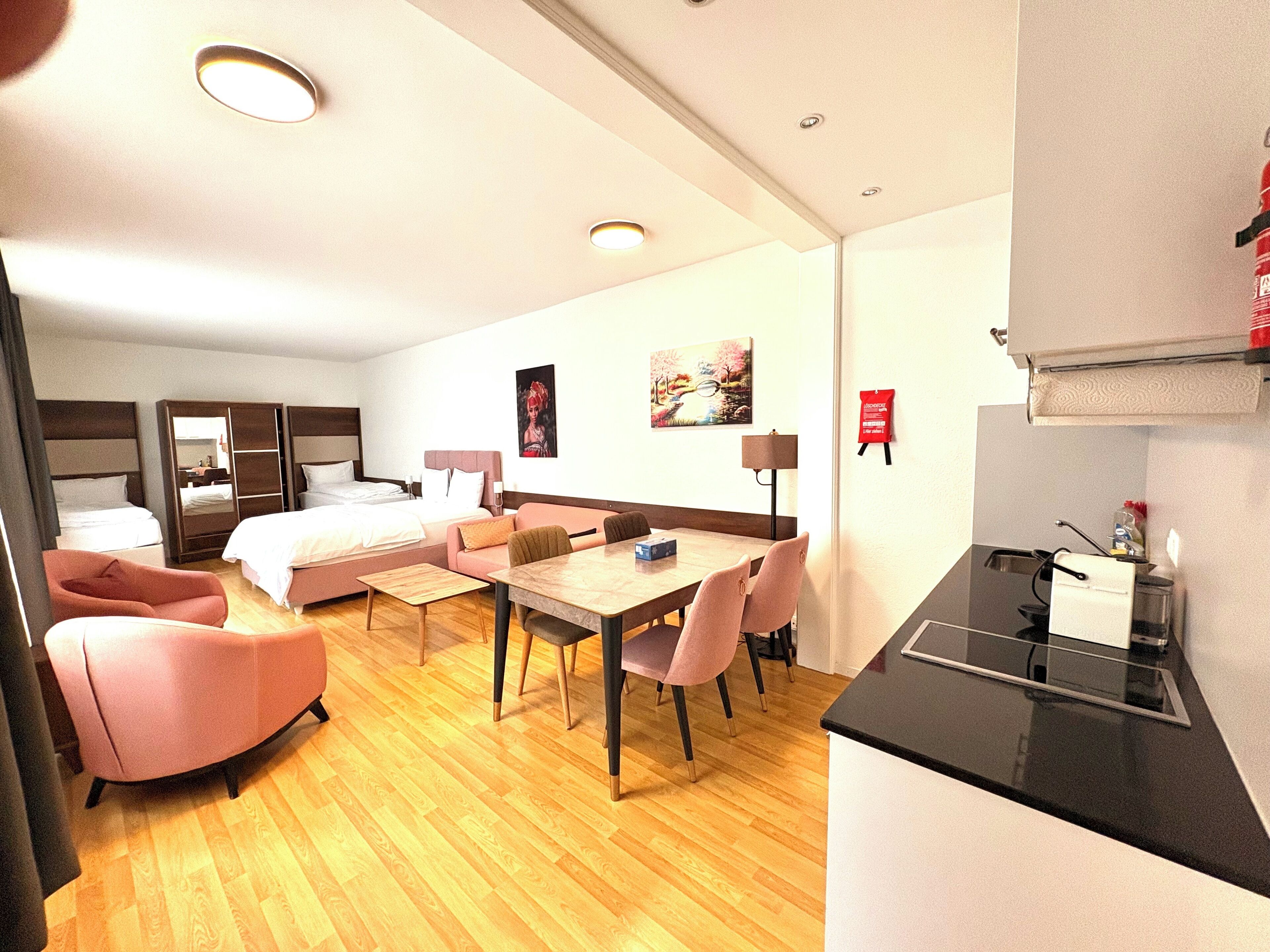 A hotel room with a bed, a sofa, a dining table with chairs, a kitchen area, and a painting on the wall.