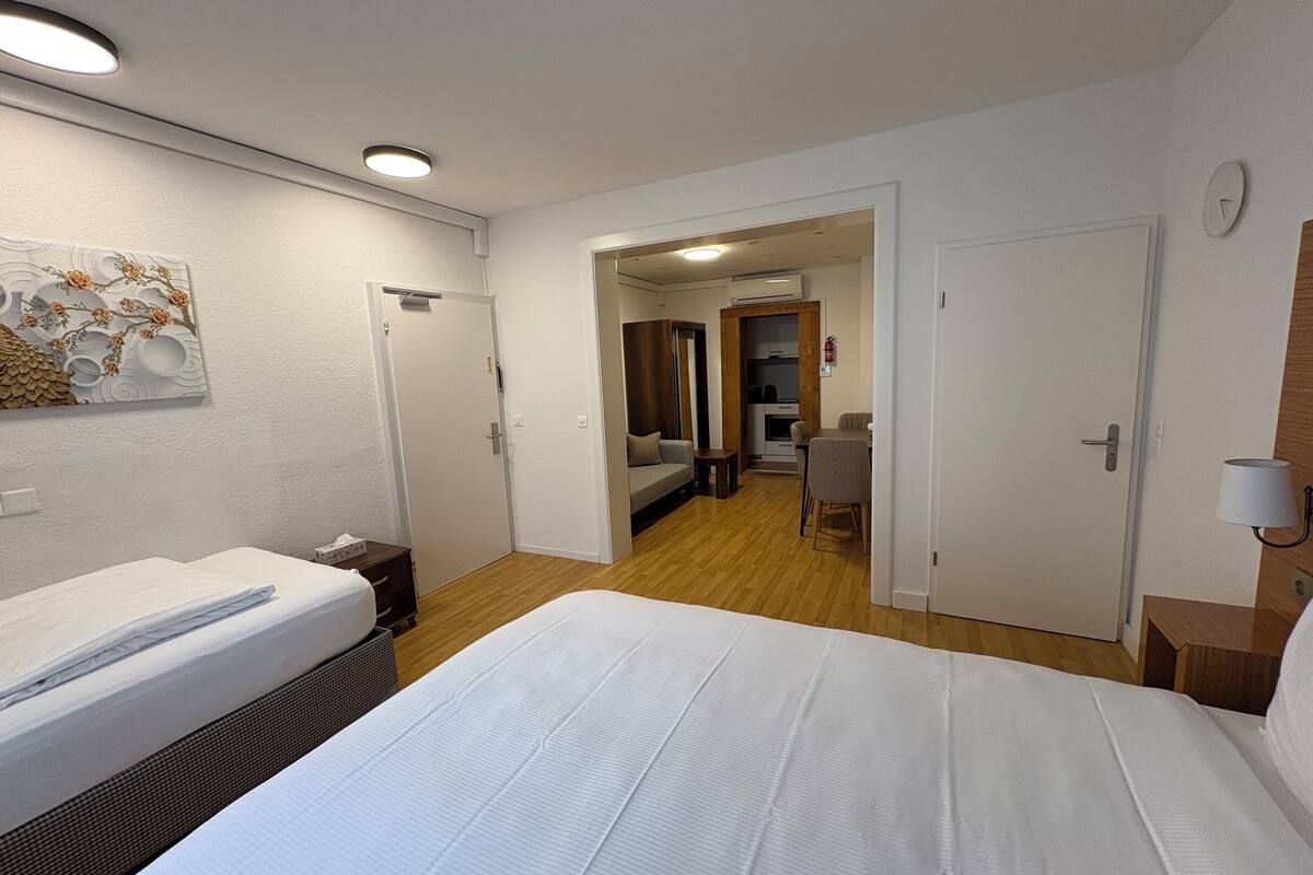 Ein Hotelzimmer mit einem großen Bett, einem Nachttisch, einem kleinen Schreibtisch und einem Sessel. Eine Tür führt zu einem Badezimmer und einem Flur.