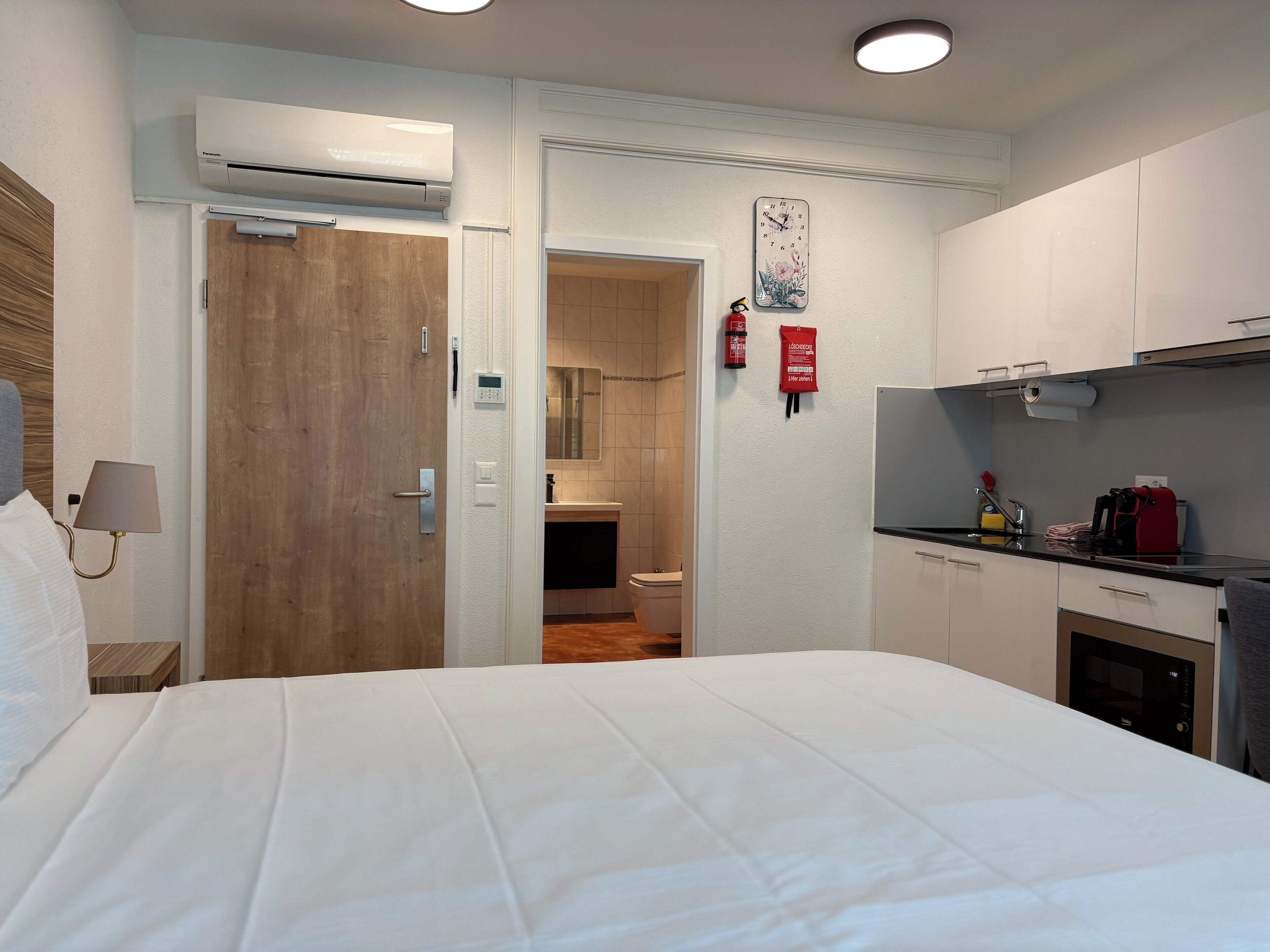 Une chambre d’hôtel comprenant un lit, une salle de bain, un coin cuisine et une horloge fixée au mur.