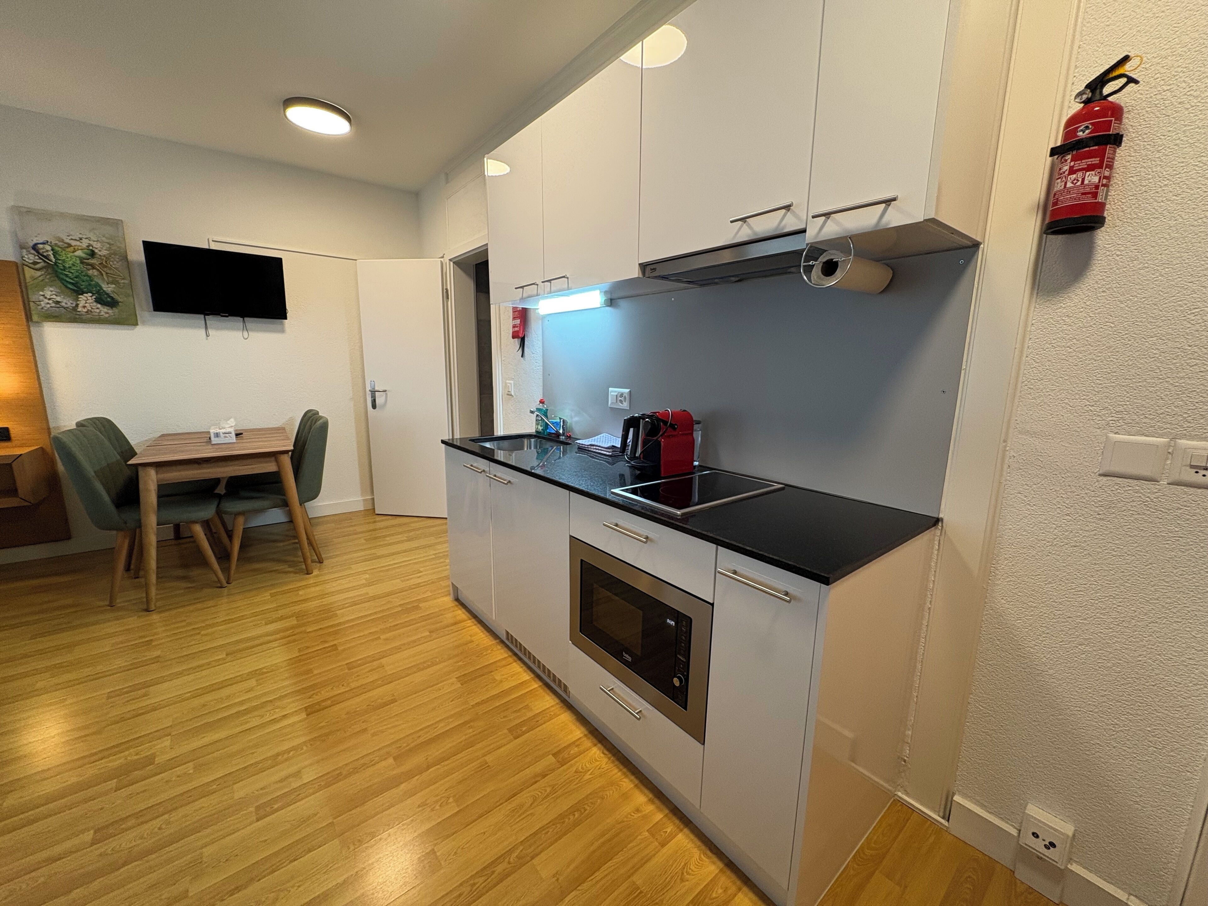 A compact kitchen with a microwave, stove, and sink. There is a dining table with chairs and a flat-screen TV on the wall.