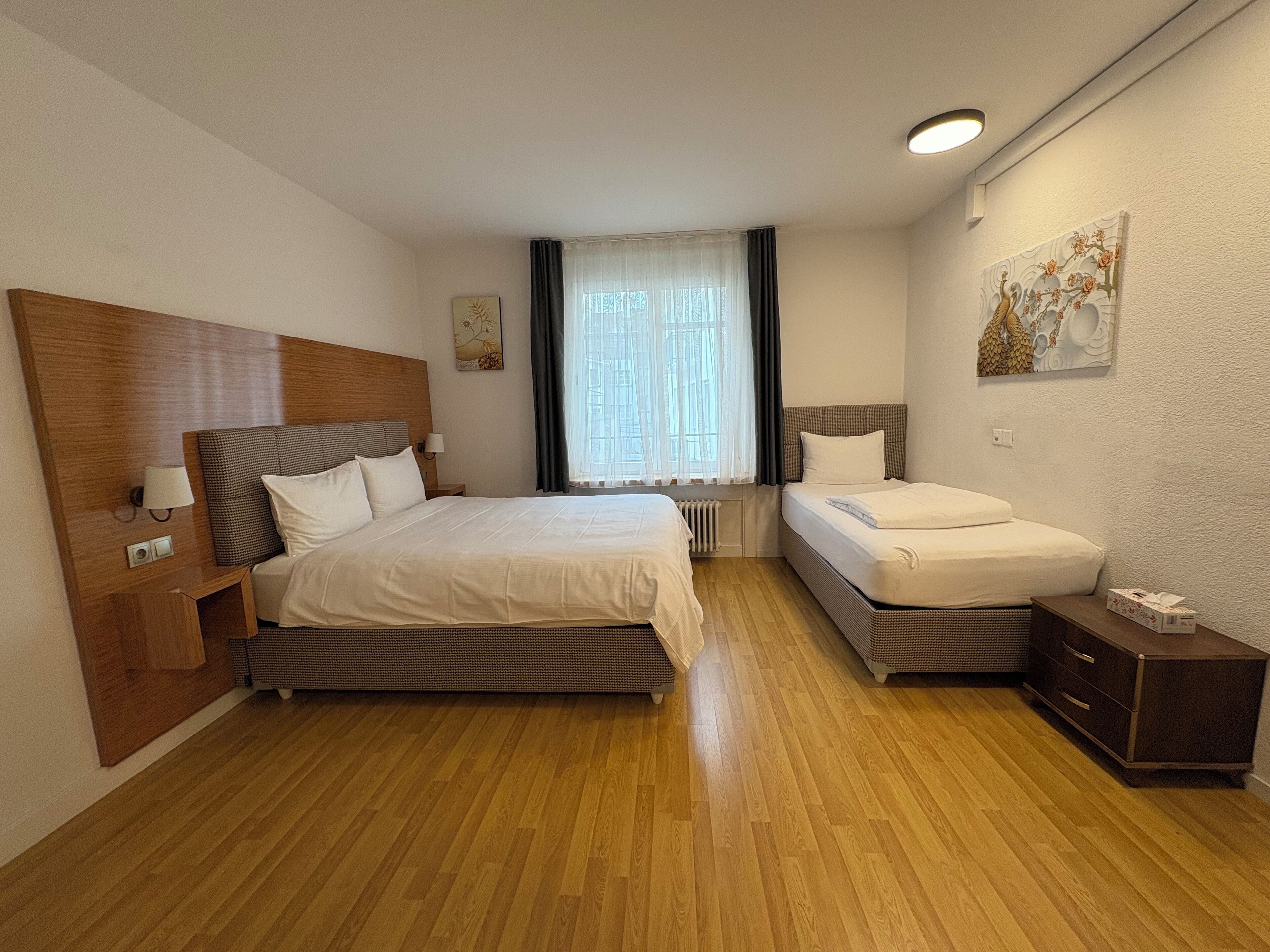 Habitación de hotel con una cama grande, una cama más pequeña, una mesita de noche, un aparador, una ventana con cortinas y una obra de arte colgada en la pared.