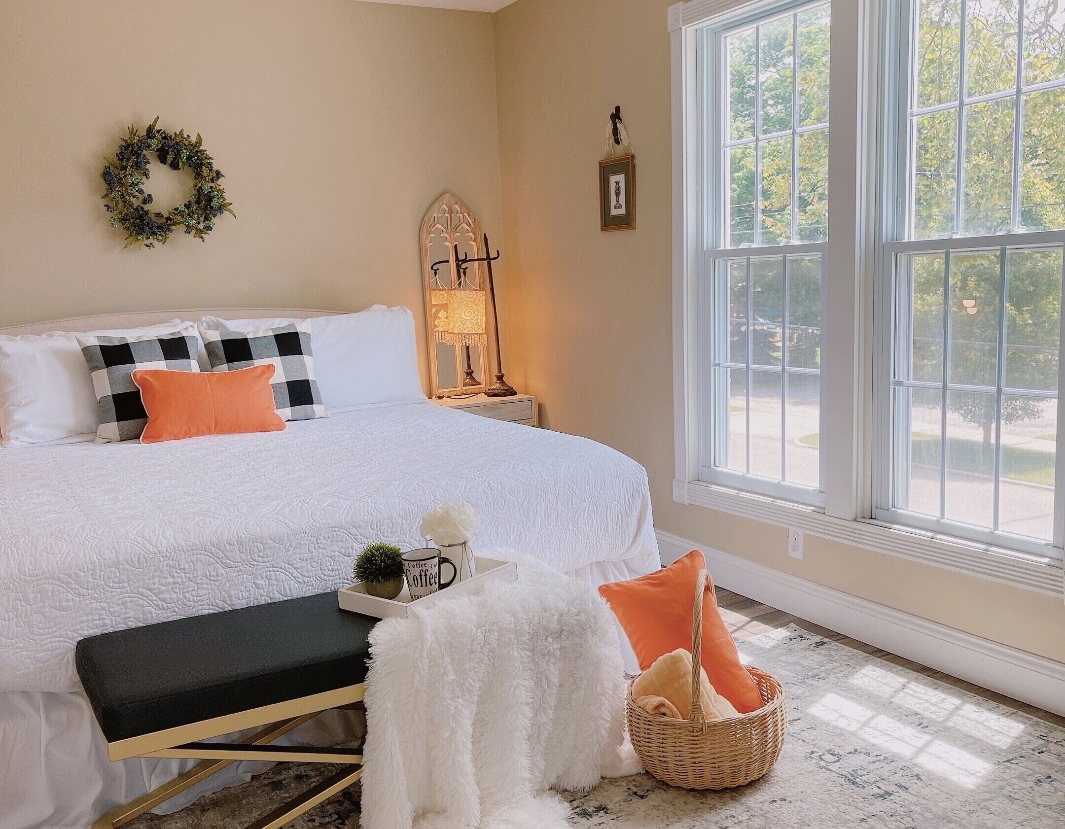 A neatly arranged bedroom with a large bed, a bedside table, a lamp, a wicker basket, and a view of the outdoors through a large window.