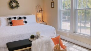 A neatly arranged bedroom with a large bed, a bedside table, a lamp, a wicker basket, and a view of the outdoors through a large window.