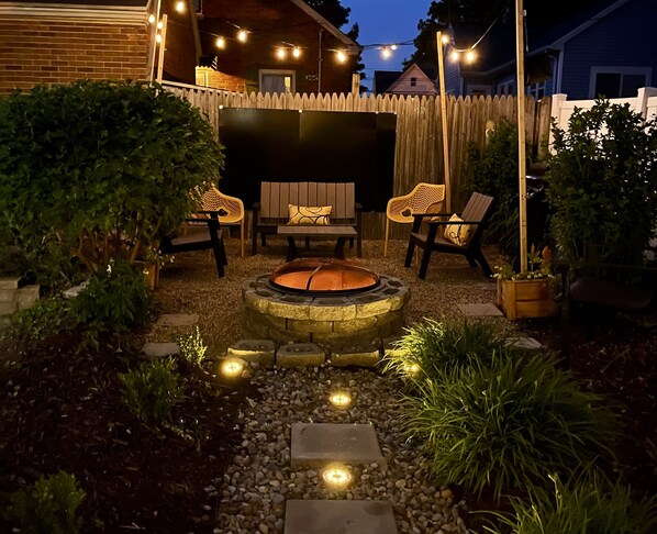 A backyard with a stone fire pit, surrounded by a wooden fence, and featuring outdoor seating with cushions.