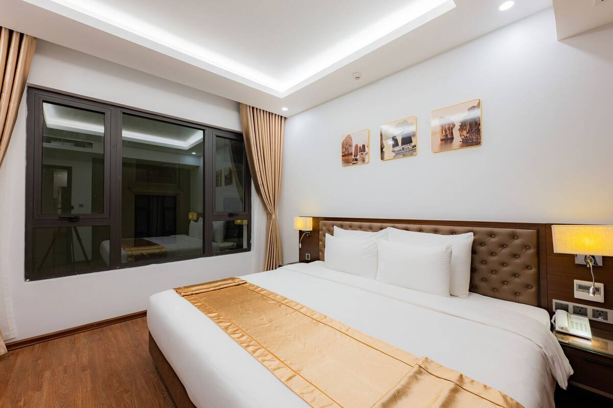 Een hotelkamer met een groot bed, een raam met gordijnen, een wandlamp en drie ingekaderde foto’s.