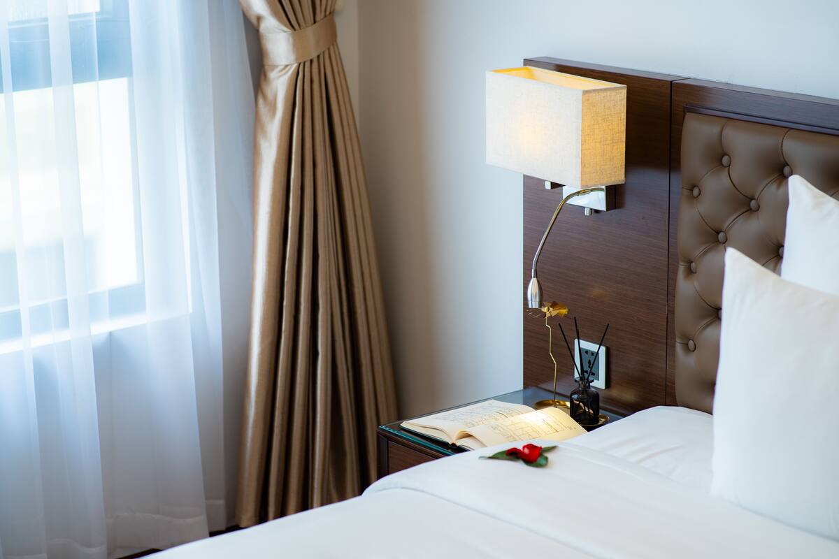 Een hotelkamer met een bed, een nachtkastje met een lamp en een raam met gordijnen.