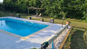 A rectangular swimming pool with a concrete edge, surrounded by a pebble border. There are lounge chairs under a pergola and potted plants around the pool area.