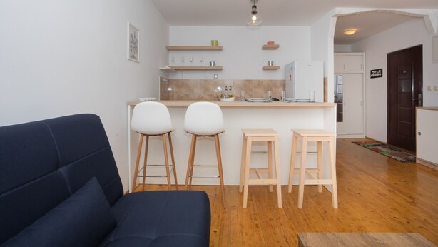 Ein modernes Wohnzimmer mit einem dunklen Sofa und drei hölzernen Hocker, eine Küche mit weißen Schränken und einer Holz Arbeitsplatte sowie ein Holzboden.