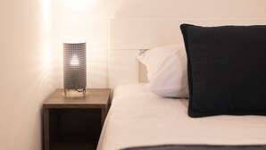 Ein ordentlich bezogenes Bett mit einem weißen und einem schwarzen Kopfkissen, ein kleiner Nachttisch mit einer Lampe und ein Holzboden.