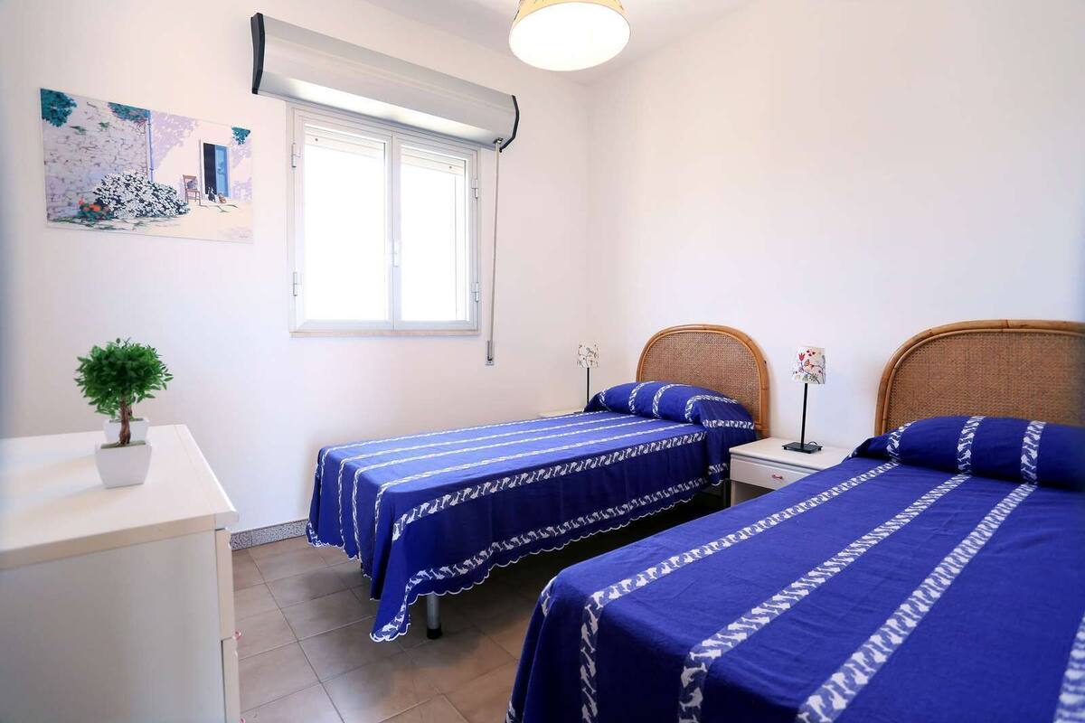 Un dormitorio con dos camas individuales, una mesita de noche pequeña, una lámpara de pared y una planta en maceta. Se aprecia una ventana con persianas.