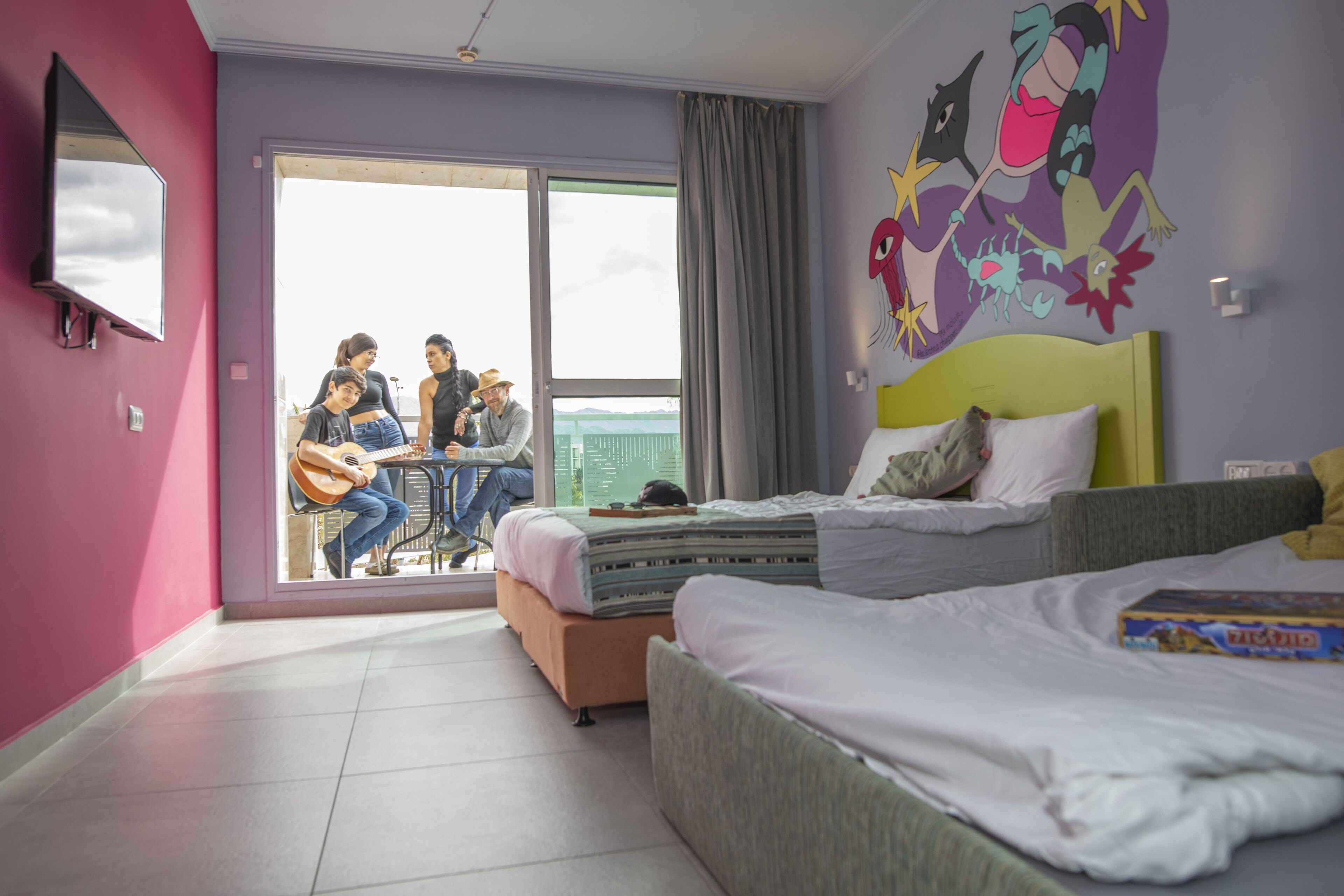 A modern hotel room with a large sliding glass door leading to a balcony, a bed with a green headboard, and a colorful mural on the wall.