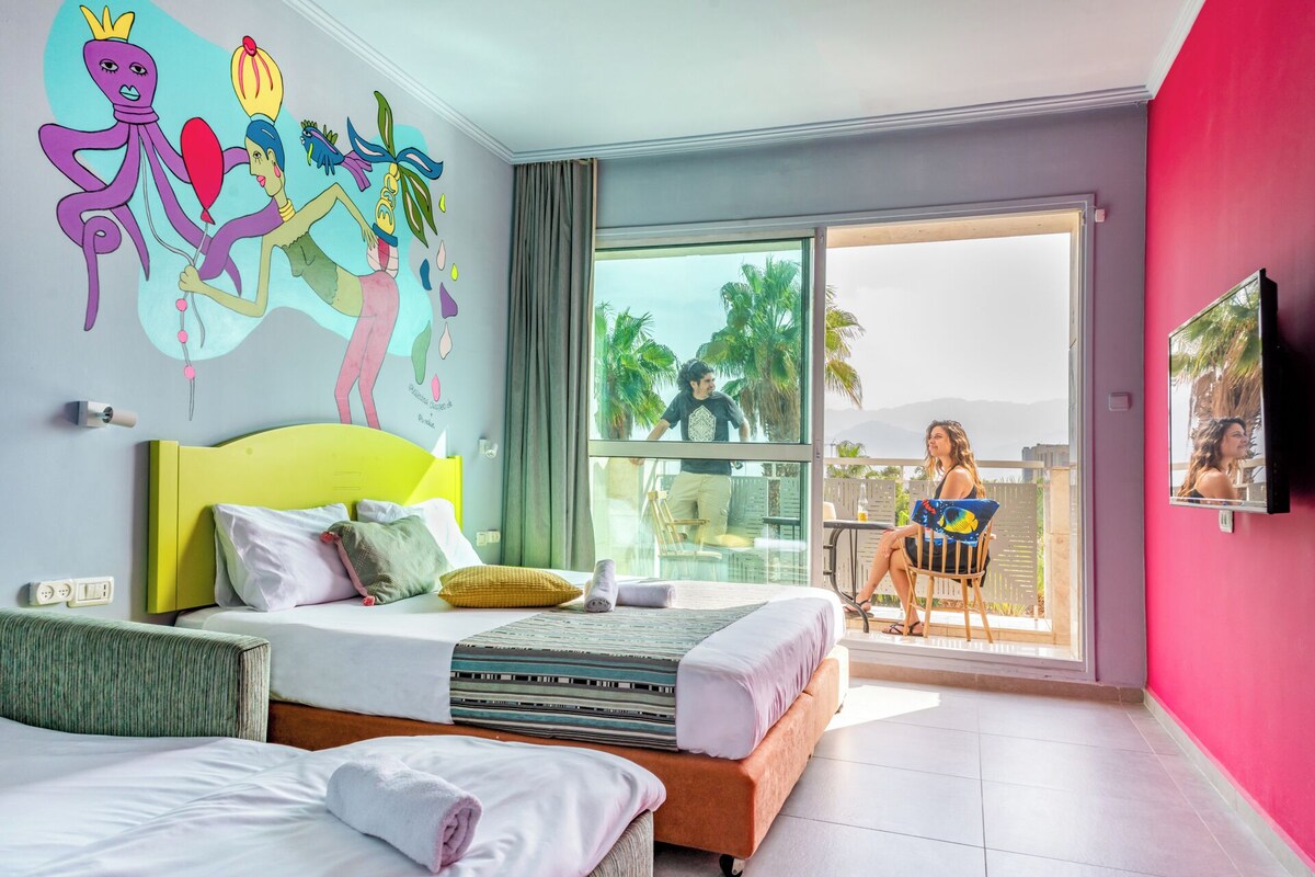 A hotel room with a large sliding glass door to a balcony, a bed with multiple pillows, a small table, and a TV mounted on the wall.