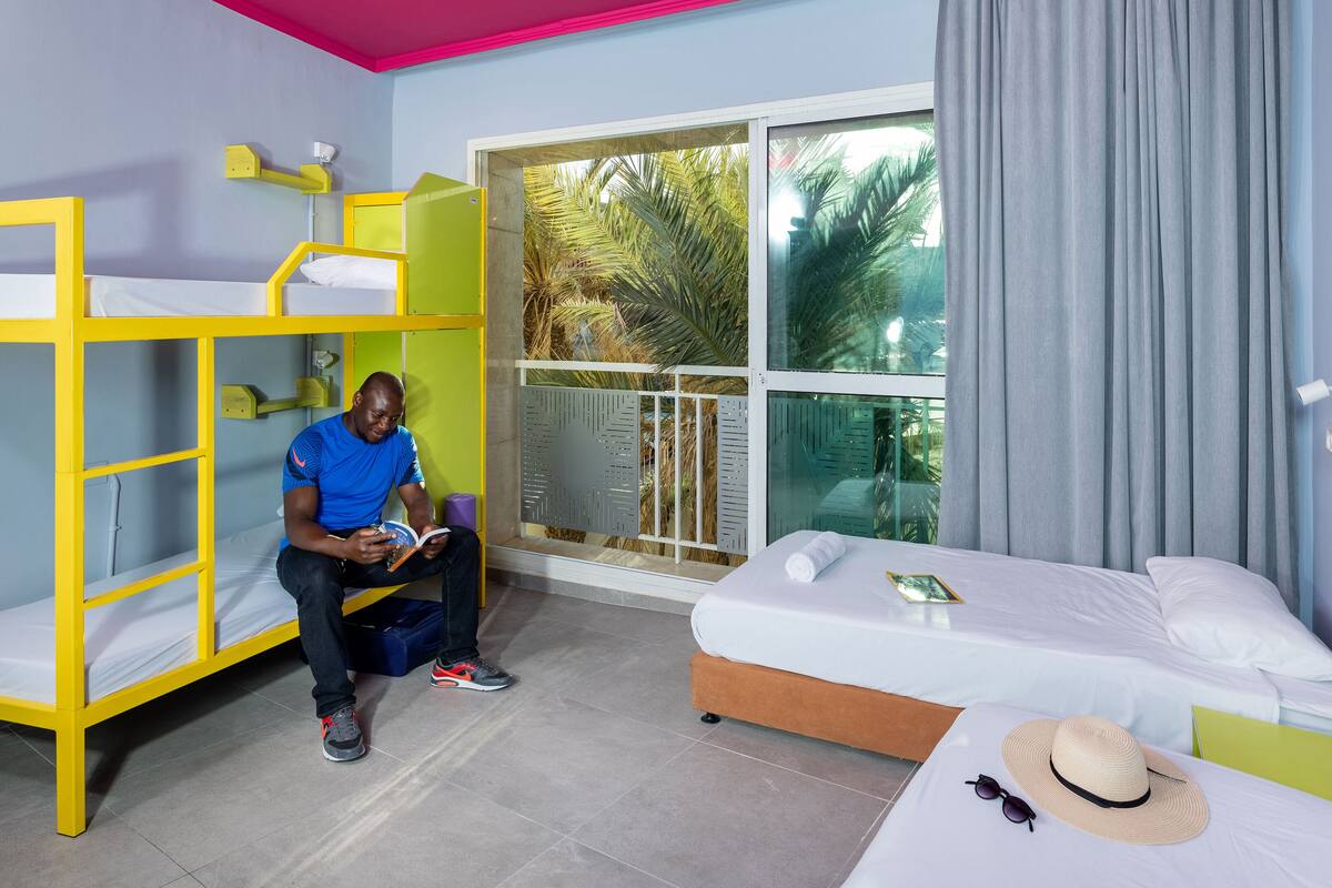 A room with bunk beds, a bed, a window with a view of palm trees, and a curtain.
