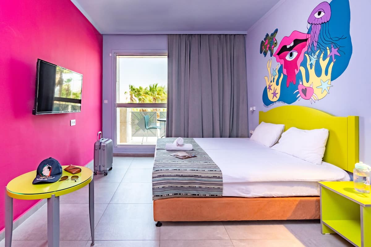A hotel room with a bed, a TV, a table with a cap and sunglasses, a suitcase, and a colorful mural on the wall.
