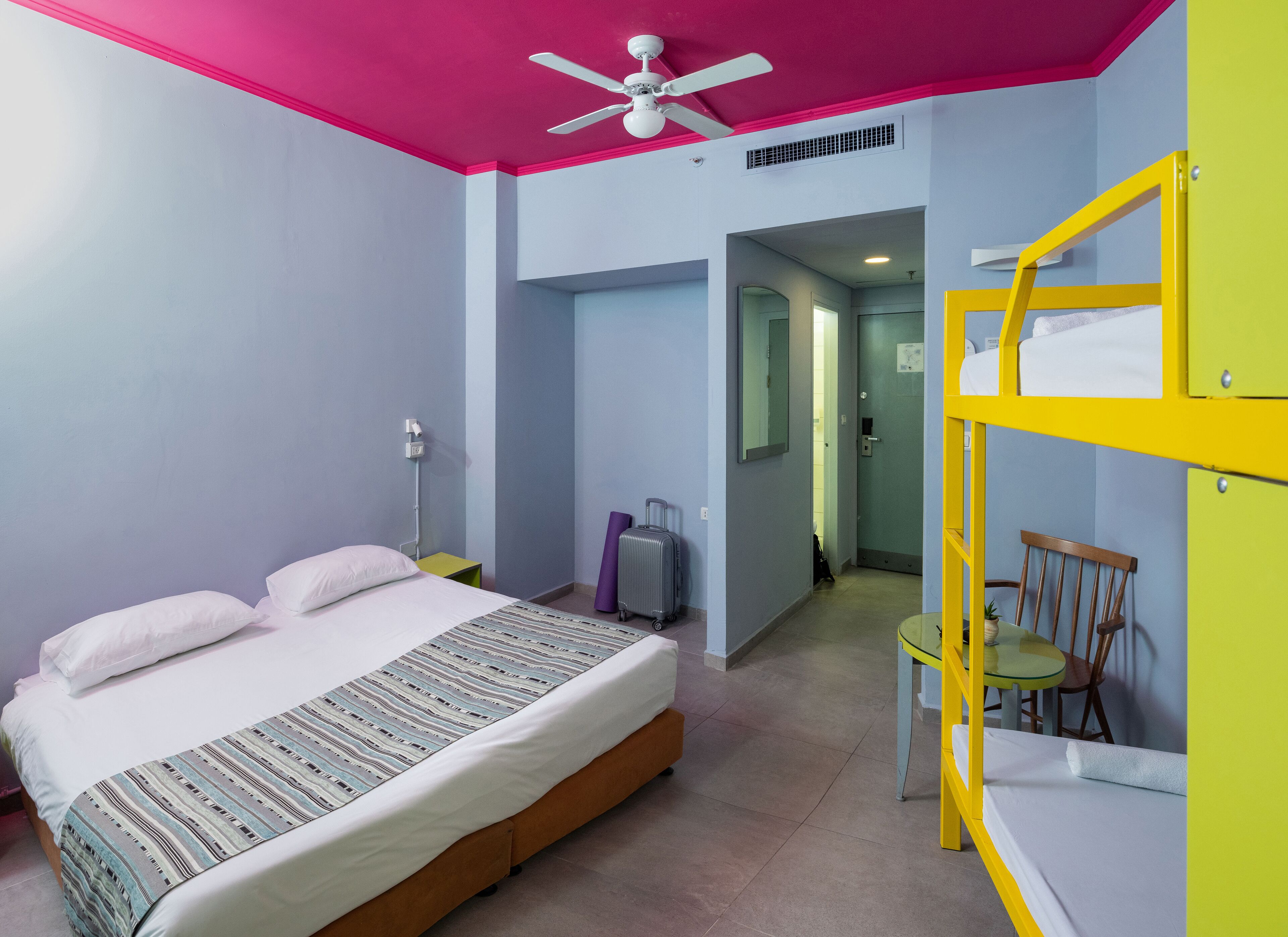 A hotel room with a bed, a ceiling fan, a bunk bed, a suitcase, and a chair.