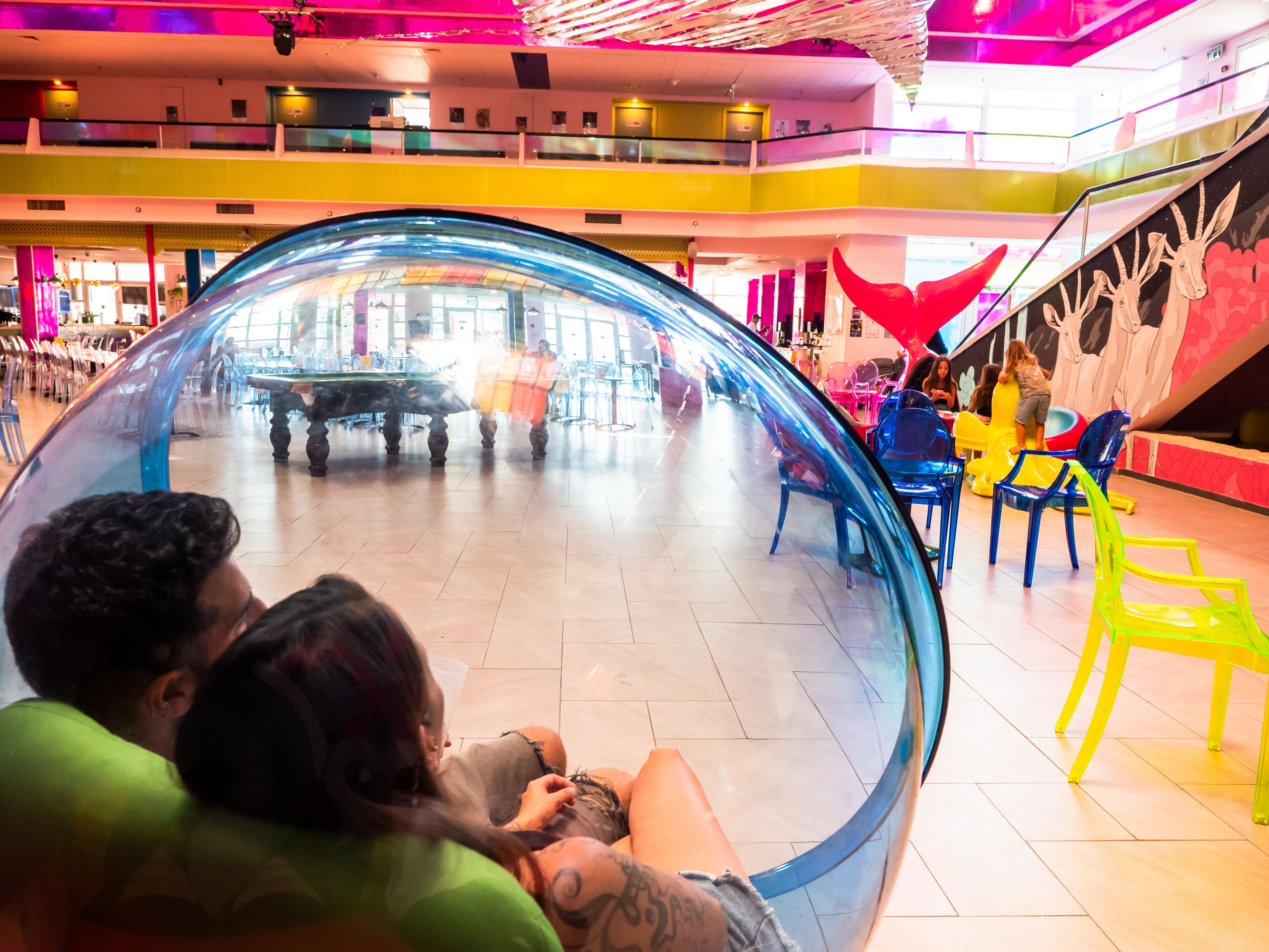 Una gran esfera transparente en un espacio público, con piso de baldosas y sillas de colores alrededor.