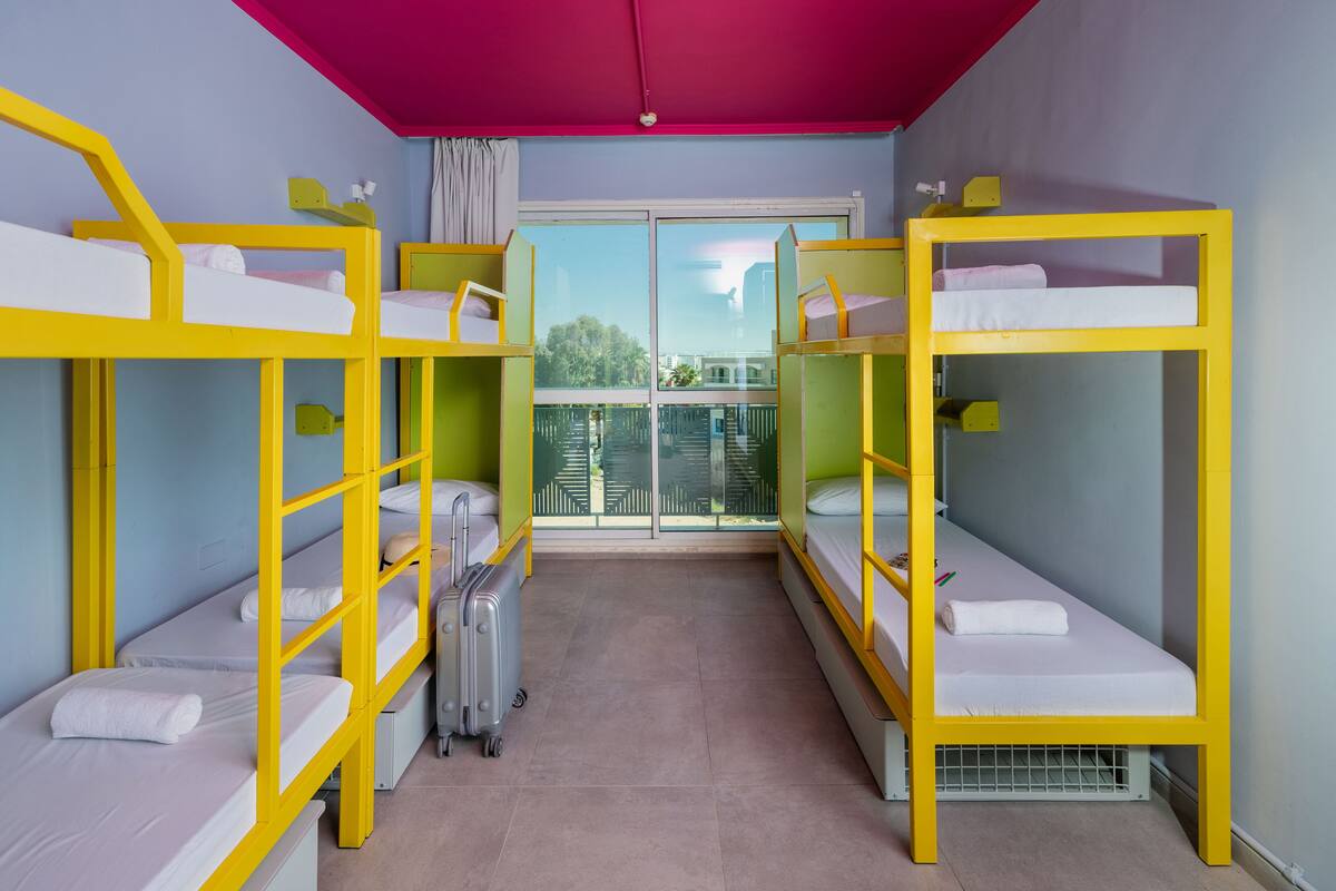 A room with bunk beds, a suitcase, and a window with a view of buildings.