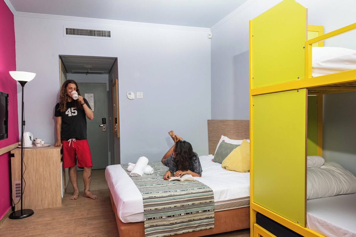 A room with a bunk bed, a desk with a kettle, and a person reading a book.