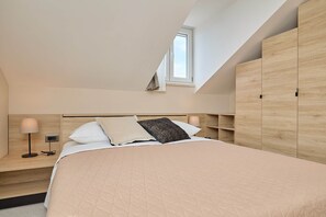 A bedroom with a sloped ceiling, a bed with a quilted bedspread, bedside lamps, and built-in wooden storage cabinets. - Hedera Estate, Hedera A32 (Dubrovnik)