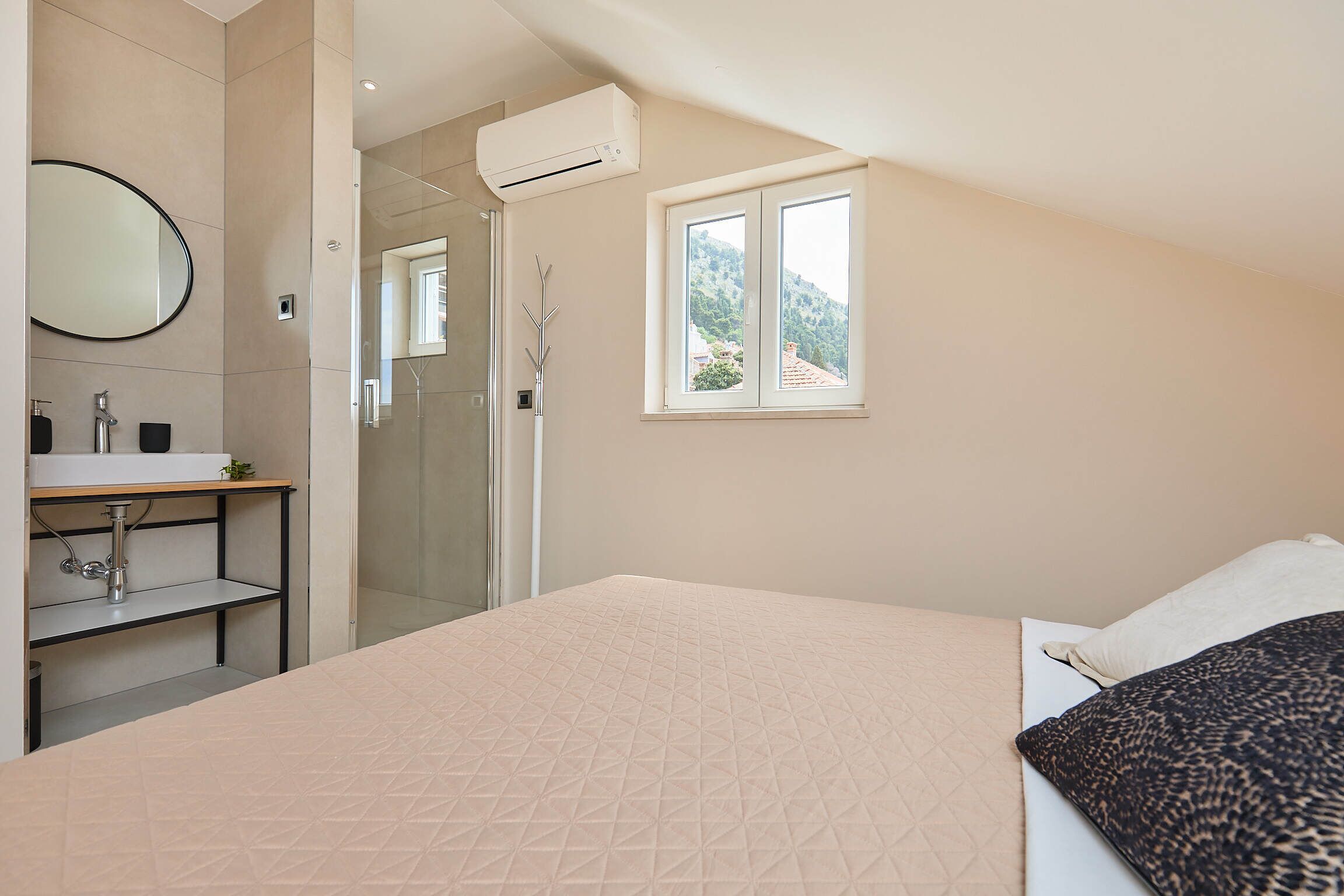 Ein Schlafzimmer mit Bett, Fenster mit Blick, Badezimmer mit Waschbecken und Spiegel sowie Klimaanlage.