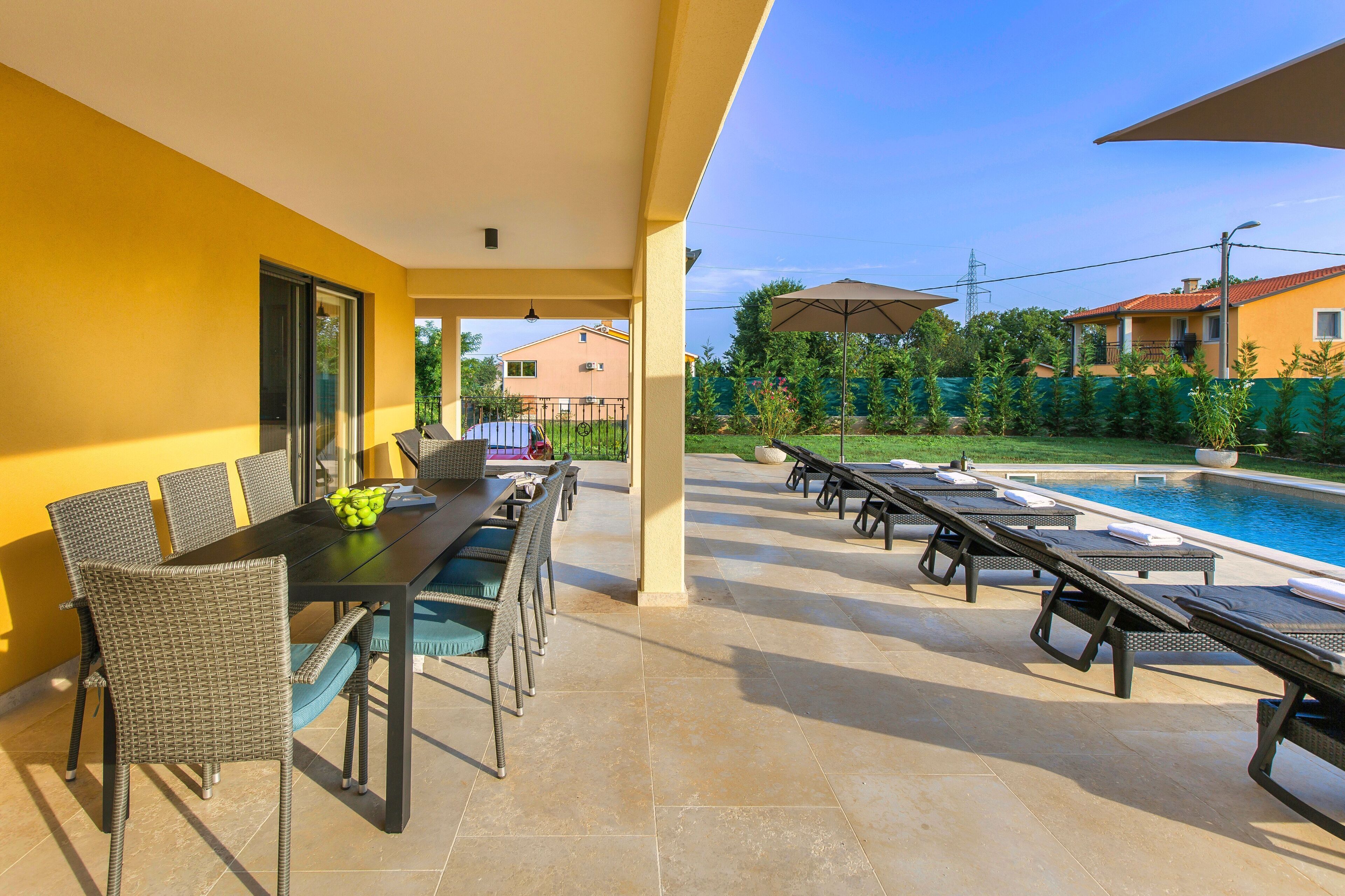 Uma área de pátio ao ar livre com mesa e cadeiras, um espaço de descanso à beira da piscina e vista para um prédio residencial ao fundo.