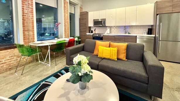 A modern living room with a grey sofa, a coffee table with a plant, and a dining area with green chairs.