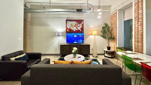 A modern living room with a sofa, a coffee table, and a TV. There is a plant, a lamp, and a dining table with chairs.
