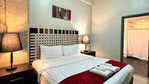 A neatly made bed with white linens and a red throw blanket, a headboard with a grid pattern, two bedside lamps, and a painting on the wall.