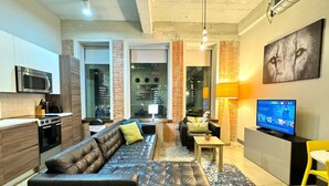 A modern living room with a leather sofa, a television, and a kitchen area with a microwave and stove. - 2 BR | 2 Bath | Downtown | Free Valet Parking | 5 (Dallas)