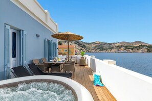 A sunlit deck with a hot tub, lounge chairs, and a table with umbrellas overlooking a coastal town. - Rementzo Apartment (Symi)