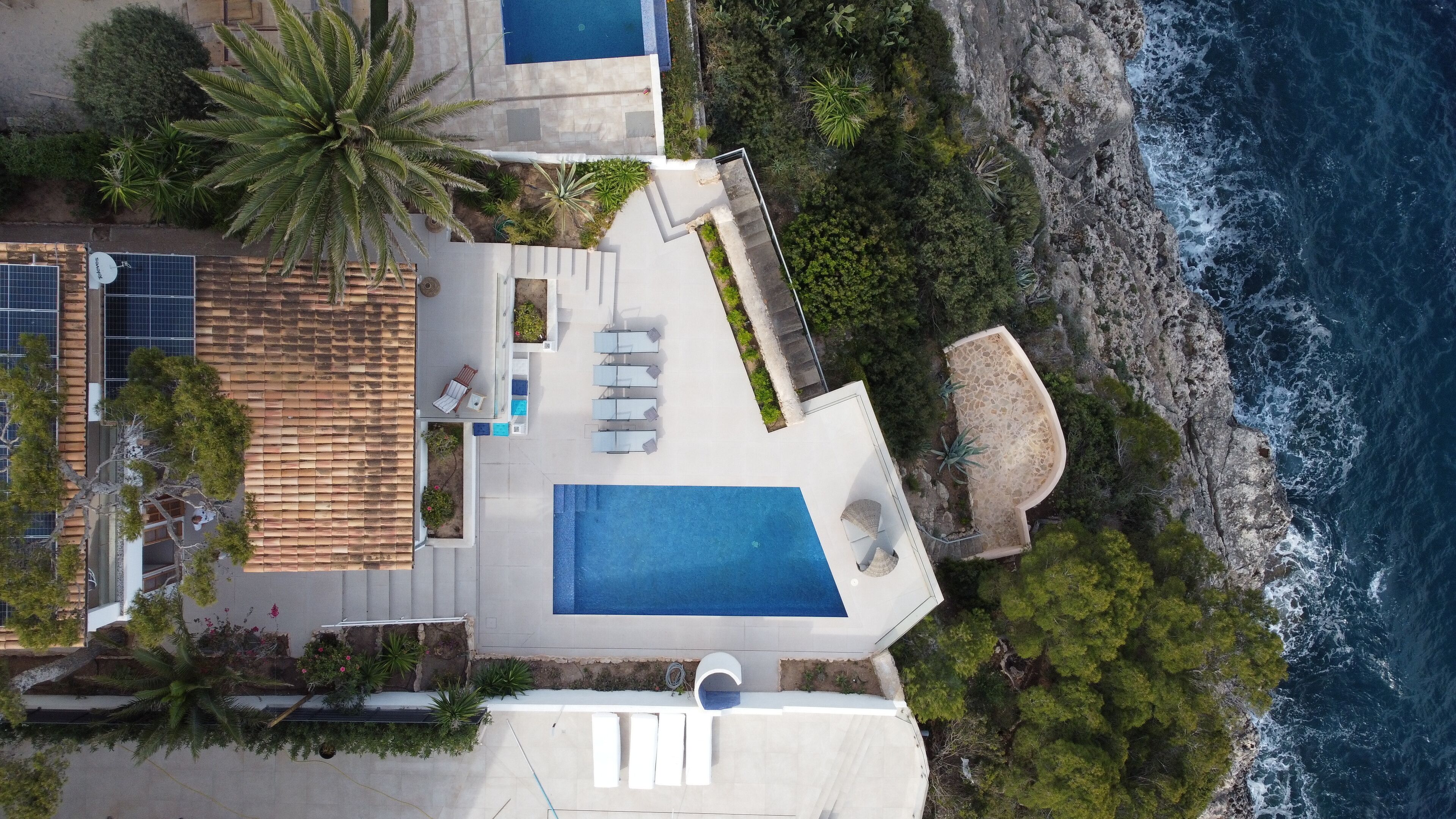 An aerial view of a coastal property with a large swimming pool, outdoor seating, and a rocky shoreline.