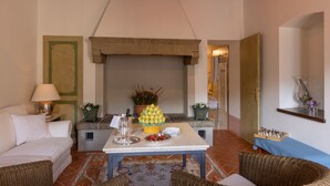 A cozy living room with a fireplace, a dining table with a fruit centerpiece, wicker furniture, and a chessboard on a shelf.
