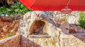 Zona de parrilla al aire libre de piedra con chimenea, rodeada de muros de piedra y vegetación.