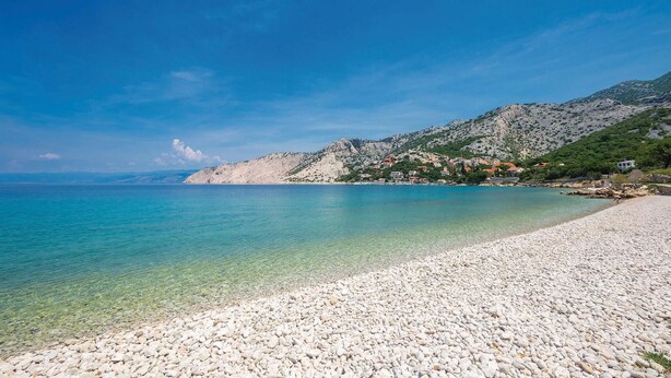 Una playa de guijarros con agua turquesa cristalina, un promontorio rocoso y un pequeño pueblo con casas de tejados rojos.